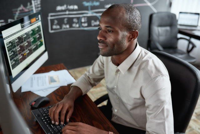 Cheerful african male trader sitting by desk and studying analytical reports using pc in the office. Stock trading, people concept. Horizontal shot