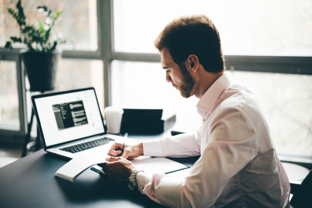 Business man working at office with laptop and documents on his desk. Man wearing white shirt and making notes on the documents.